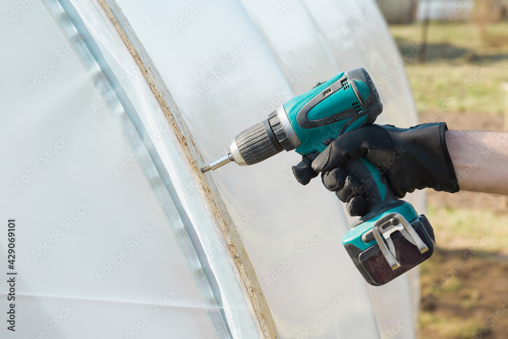 Man hand in protective glove holding battery screwdriver and assembling new polythene greenhouse. Closeup. Side view. Preparation for garden season in spring. Man hand in protective glove holding battery screwdriver and assembling new polythene greenhouse. Closeup. Side view. Preparation for garden season in spring.