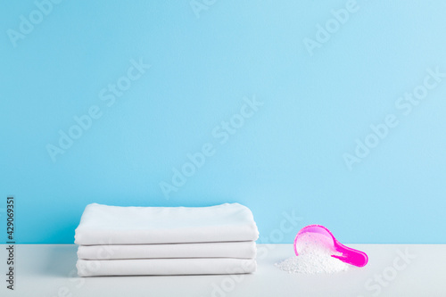 Stack of white bed sheets and cup or scoop of powder on table at light blue wall background. Detergent for clothes washing. Pastel color. Front view. Closeup. Empty place for text.  photo