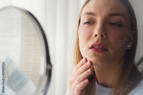 Young beautiful woman looking herself in the mirror at home. Worried about acne caused by wearing a mask. Maskne. Problems with acne. © lordn