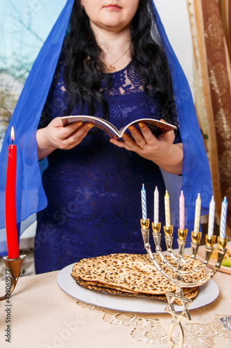 A brunette Jewish woman with her head covered in a blue cape at the Passover Seder table is reading the Passover Haggadah.