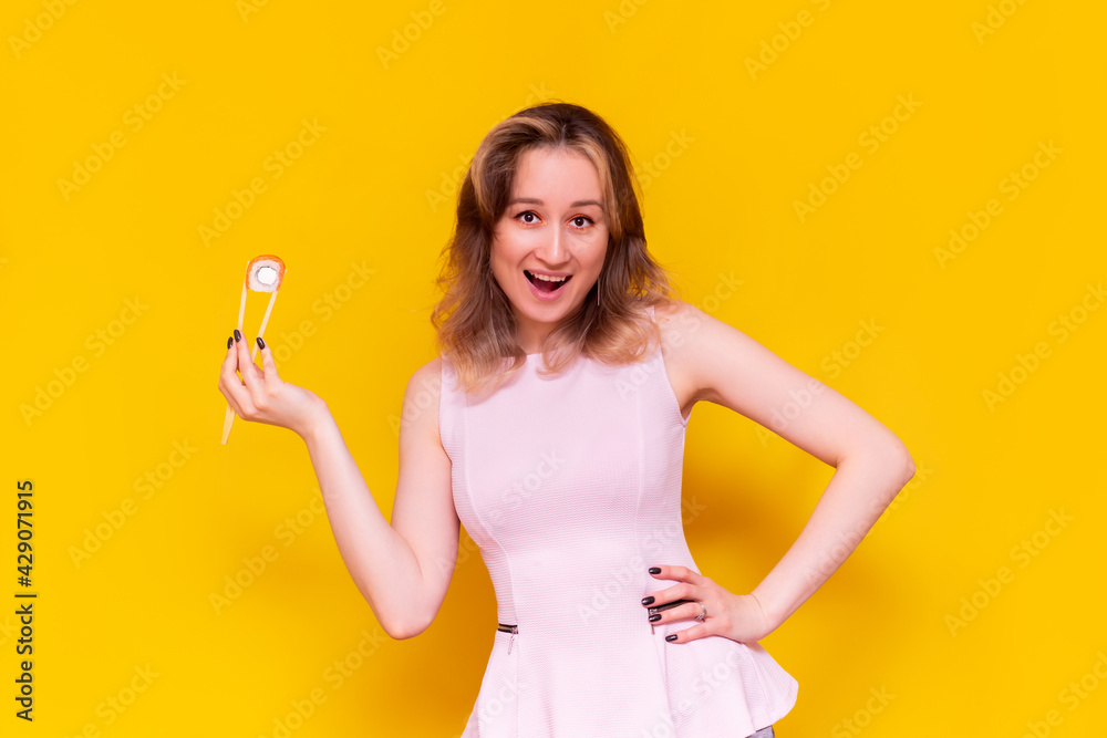 A young caucasian pretty happy excited smiling brown-haired woman in a light pink blouse advertises sushi holding in bamboo sticks roll philadelphia isolated on a bright color yellow background A young caucasian pretty happy excited smiling brown-haired woman in a light pink blouse advertises sushi holding in bamboo sticks roll philadelphia isolated on a bright color yellow background