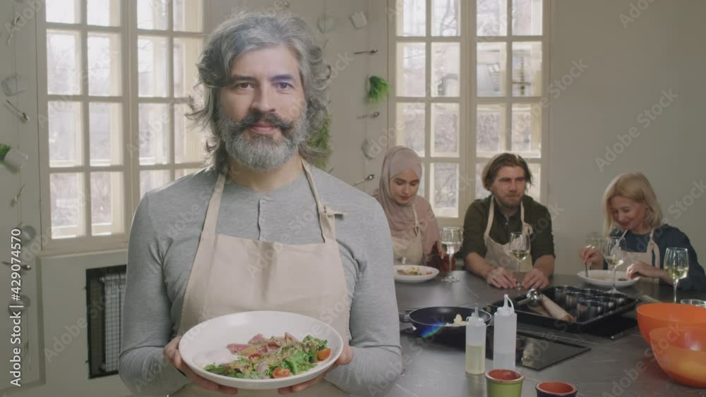 Slow-motion medium portrait of handsome adult man with grey hair and beard standing at cooking class holding just cooked dish on plate looking at camera Slow-motion medium portrait of handsome adult man with grey hair and beard standing at cooking class holding just cooked dish on plate looking at camera
