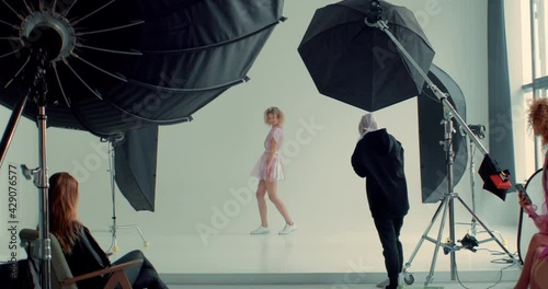 WIDE FIXED Behind the scenes of a photo shoot, team working on a fashion shot, professional female photographer taking pictures of a model in a bright studio. Shot with 2x anamorphic lens