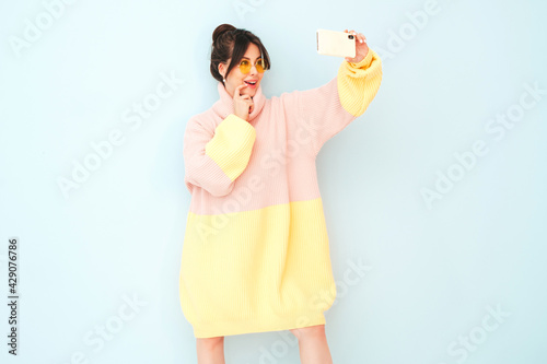 Young beautiful smiling female in trendy summer colorful sweater. Sexy carefree woman posing near light blue wall in studio. Positive model having fun indoors in sunglasses.Taking selfie