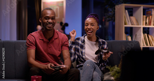 Young african beautiful happy couple playing exciting video game on console using gamepads having fun in living room. Apartment. Entertainment.