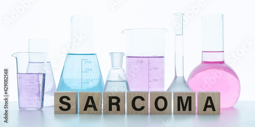 Wooden block form the word SARCOMA. In the background are flasks with multi-colored liquids. Medical concept.