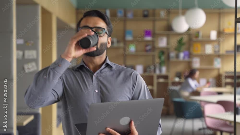Happy indian businessman holding coffee and laptop walking in office. Smiling male employee, business man executive going along modern office coworking space using computer.
