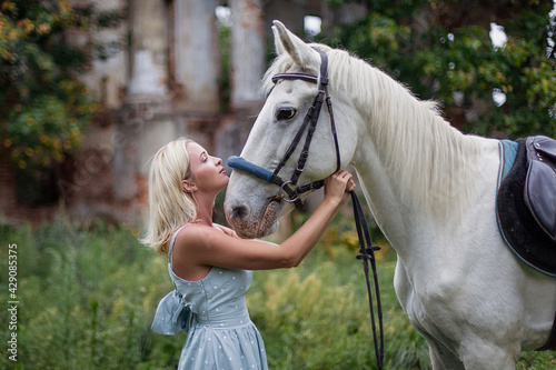 Beautiful woman in a blue dress hugs a white horse. Best friends. Photoshoot with a horse. Hobby love for animals. Country life. Romantic elegant girl tenderness. Saddle and bridle. Close-up portrait