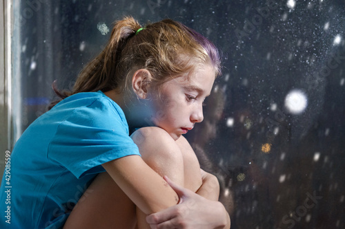 A brown eyed girl in blue t-shirt sitting on the window sill behind a foggy window with snowy night background