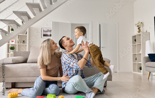 Cheerful young couple having fun with happy cute kid boy relaxing in modern house living room. Parents playing with funny toddler child son, bonding together spending time sitting on floor at home.