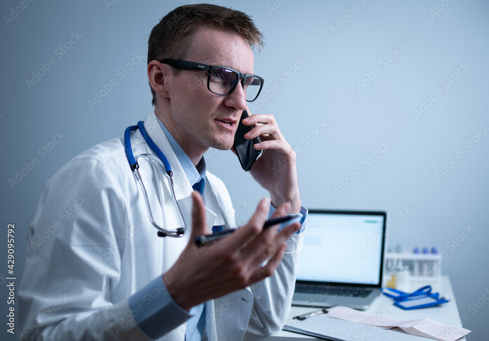 European doctor in white medical coat and glasses consults patient on cell phone in clinic office, sitting at table with laptop. German general practitioner at workplace in hospital talking on phone European doctor in white medical coat and glasses consults patient on cell phone in clinic office, sitting at table with laptop. German general practitioner at workplace in hospital talking on phone