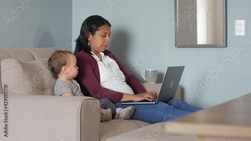 A Hispanic mother dressed in red and white is sitting in a couch working or studying online at a laptop computer together with a baby girl 