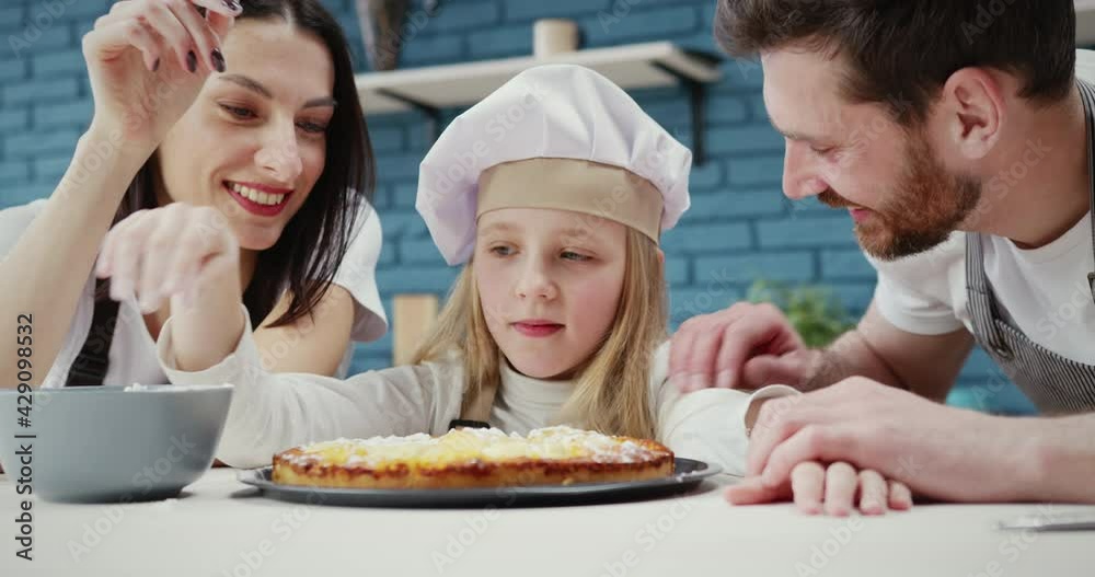Happy smiling mum and dad helping excited little girl daughter to sprinkle powdered sugar on homemade pie. Family members feeling satisfied with their work in kitchen. Happy smiling mum and dad helping excited little girl daughter to sprinkle powdered sugar on homemade pie. Family members feeling satisfied with their work in kitchen.