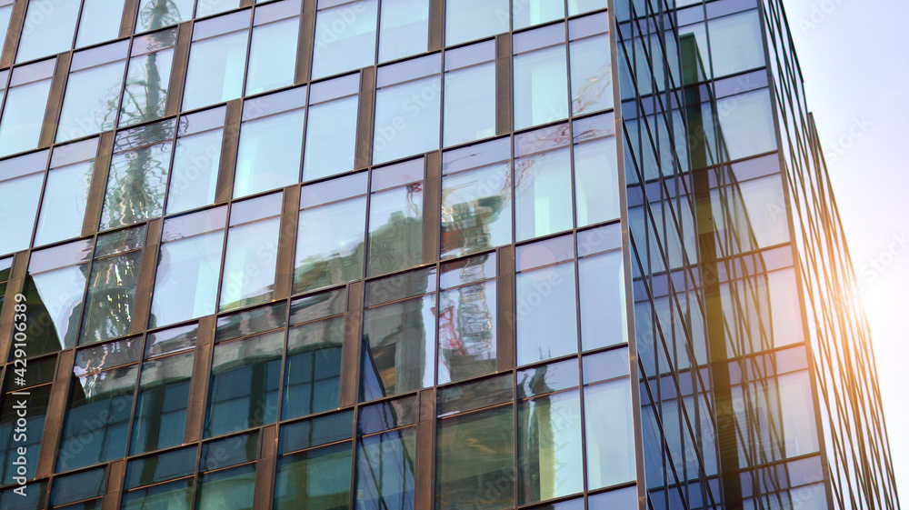 Obraz premium Blue sky reflection in glass facade of building. View of office building windows close up with sunrise, reflection and perspective.. Glass facade on a bright sunny day with sunbeams on the blue sky. 