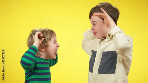 Side view portrait of playful carefree Caucasian brothers or friends making faces with tongue out at yellow background. Cheerful boys having fun grimacing looking at each other