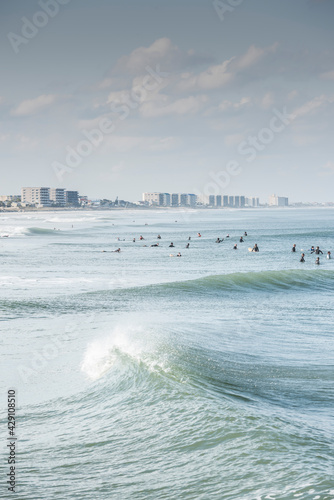 Photography Many surfers waiting in a line up to catch a wave as a swell rolls by with the Daytona Beach, Florida skyline in the background
