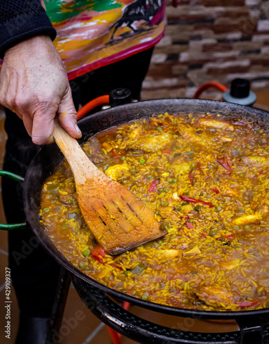 Chef hand with a wooden spatula cooking paella. Chef cooking Spanish traditional recipe. Mediterranean paella with red pepper, shrimps, peas and chicken. Yellow rice texture. Vertical shot paella.