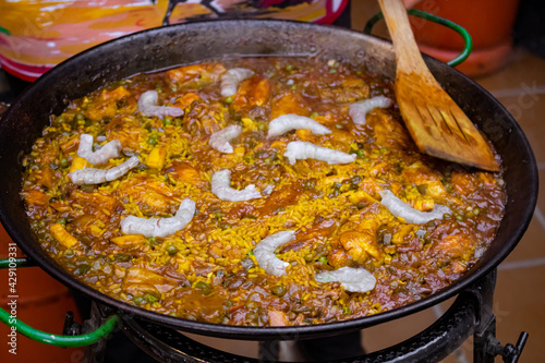 Chef with a wooden spatula cooking paella in a paella pan. Chef cooking Spanish traditional recipe. Mediterranean paella with red pepper, shrimps, peas and chicken. Yellow rice texture.
