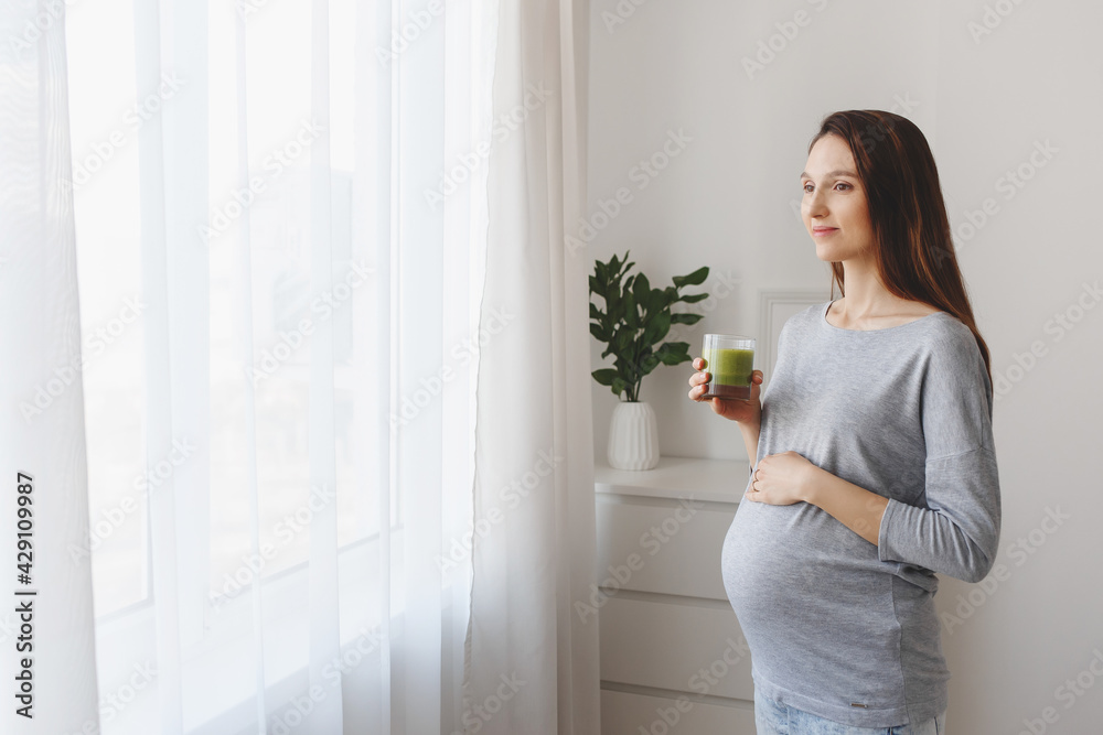 Young beautiful pregnant woman standing at home near the window and drinking a green smoothie. Young beautiful pregnant woman standing at home near the window and drinking a green smoothie.