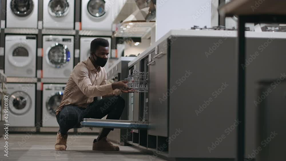 afro-american man with face mask is choosing dishwasher in trading area of modern home appliance store, opening door and examining inside afro-american man with face mask is choosing dishwasher in trading area of modern home appliance store, opening door and examining inside