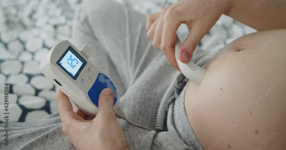 Young pregnant woman use pocket ultrasound doppler to check baby heart rate. The girl runs the sensor over her stomach, trying to detect the baby's pulse.
