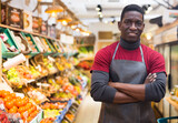 Smiling confident african american greengrocery owner wearing in black apron standing near shelves with fruits and vegetables
