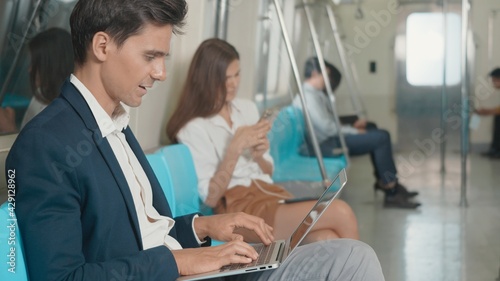 Young Caucasian white businessman sitting using laptop computer working on the subway train in the morning during going to work, people transportation concept