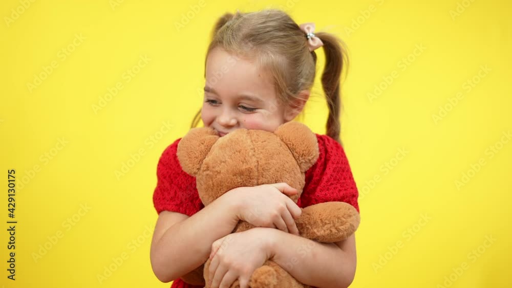 Adorable little girl hugging teddy bear standing at yellow background. Portrait of smiling happy Caucasian kid playing with toy. Childhood leisure and lifestyle