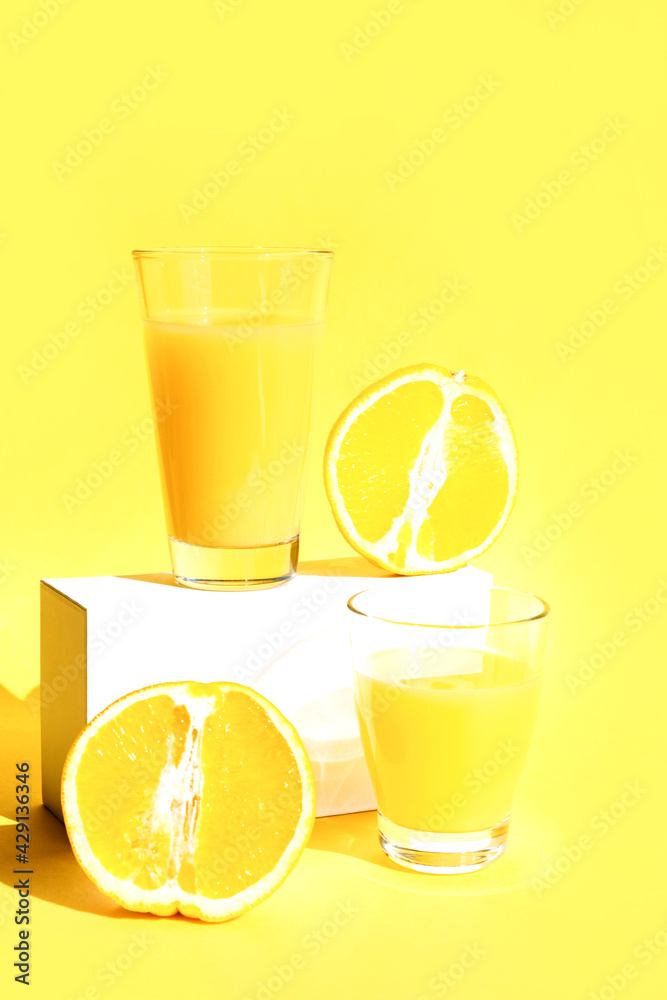 Fototapeta premium A glass of freshly squeezed refreshing orange juice on a white pedestal or podium with orange halves. Yellow background. The concept of a healthy lifestyle, raw food diet, vegetarianism. Copy space