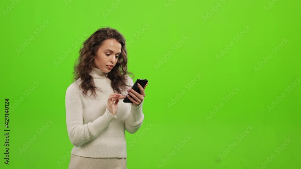 The woman is looking through social networks on her smartphone. She is standing on a green background. Green screen. 4K The woman is looking through social networks on her smartphone. She is standing on a green background. Green screen. 4K