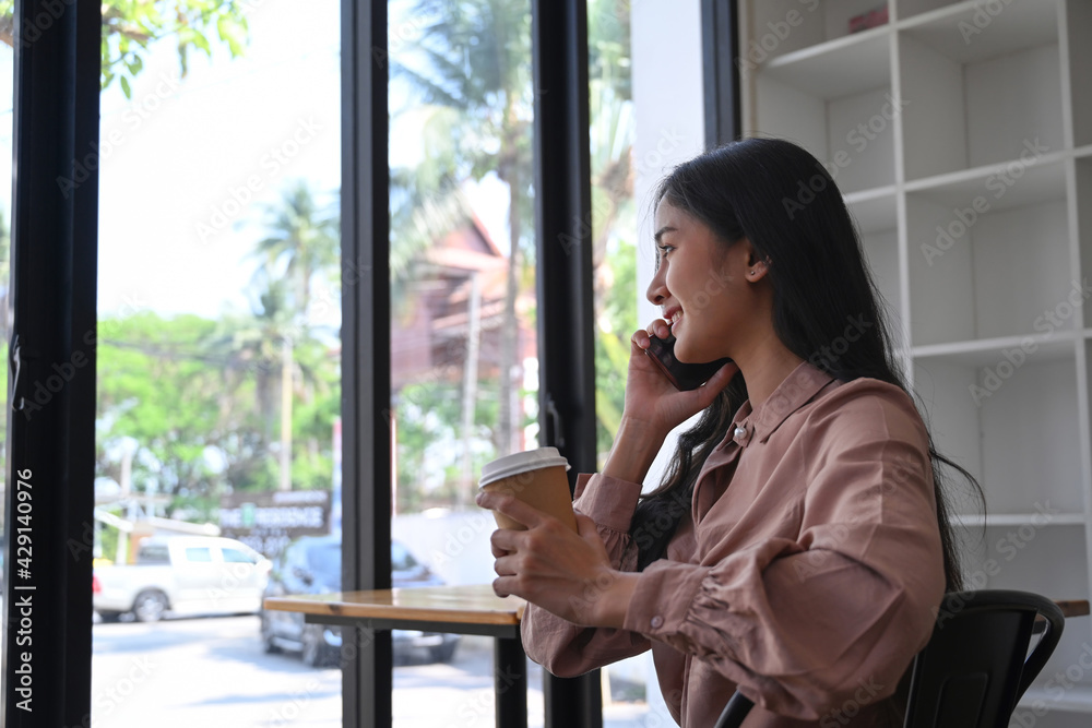 Happy young businesswoman holding coffee cup and talking on mobile phone while sitting near window in office building. Happy young businesswoman holding coffee cup and talking on mobile phone while sitting near window in office building.