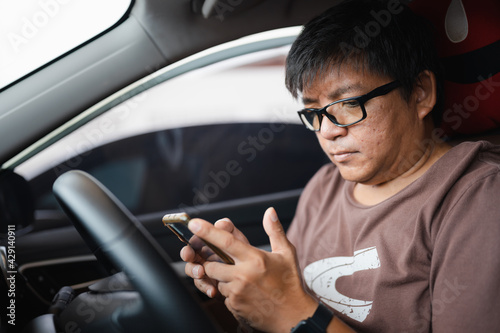 obese Asian man with eyeglasses without seatbelt looking at smartphone to check text message during traffic jam, dangerous driving, distract from attention may cause car accident © Thitiporn