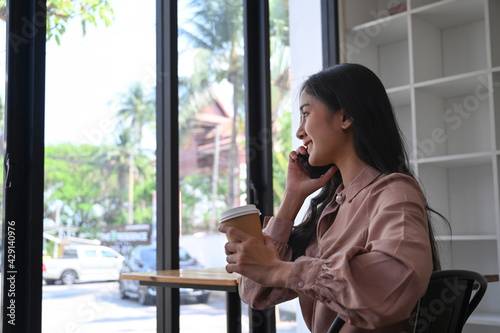 Happy young businesswoman holding coffee cup and talking on mobile phone while sitting near window in office building.