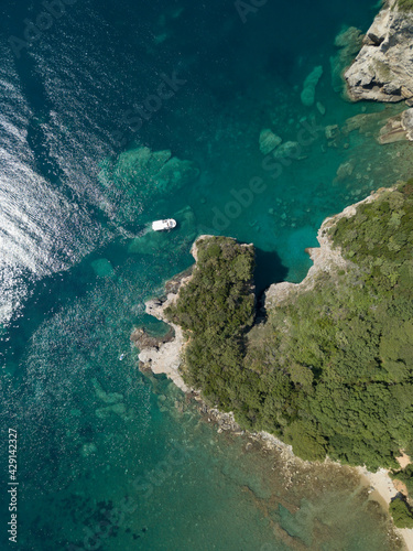Aerial view of boat sailing near steep stone cliff with green forest above transparent emerald water of Adriatic sea. Stones on seabed. Unspoiled nature of Montenegro coast. Budva scenic landscape