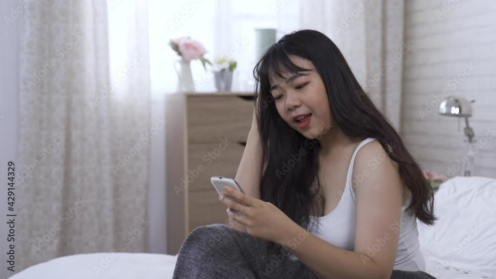 happy asian woman is touching her head while texting with smartphone on bed at background bright home interior. happy asian woman is touching her head while texting with smartphone on bed at background bright home interior.