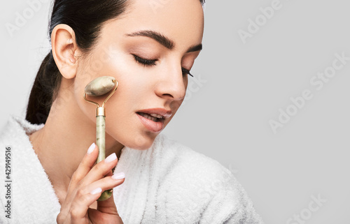 Facial jade roller. Young woman massages her face using a green jade roller for elastic perfect skin. Isolated on grey