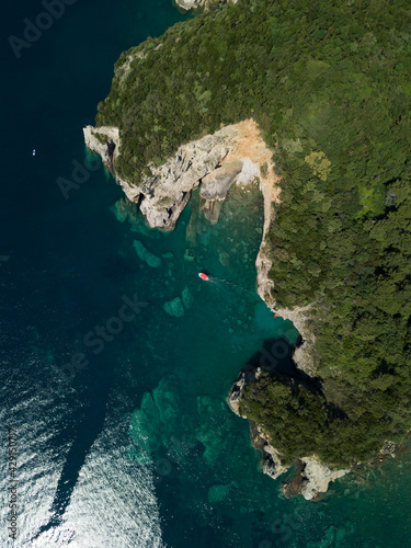 Aerial view of red boat sailing near steep stone cliff with green forest above transparent emerald water of Adriatic sea. Stones on seabed. Unspoiled nature of Montenegro coast. Budva scenic landscape