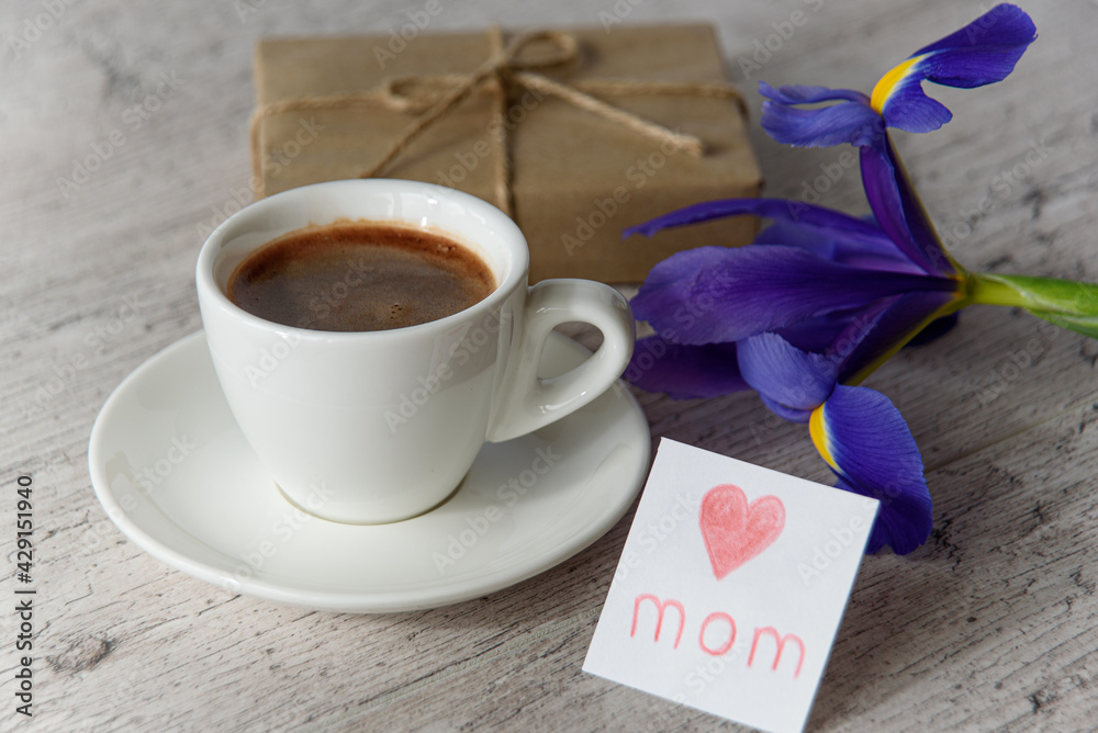 White cup of coffee, card for mom from child, gift box and iris flower on gray wood table. Happy Mothers Day.