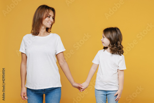 Happy woman in basic white t-shirt have fun with child baby girl 5-6 years old hold hands. Mom little kid daughter isolated on yellow orange color background studio. Mother's Day love family concept.
