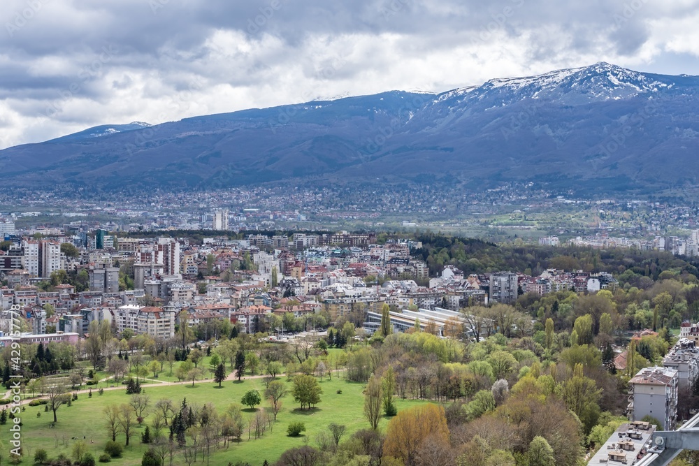 Aerial view of urban landscape with many administrative and residential buildings, green park and mountain background, cloudy sky. Sofia, the capital city of Bulgaria, East Europe. Panoramic sight.
