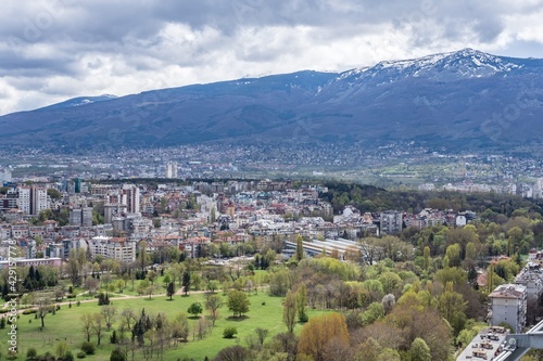 Aerial view of urban landscape with many administrative and residential buildings, green park and mountain background, cloudy sky. Sofia, the capital city of Bulgaria, East Europe. Panoramic sight. © Miglena Pencheva