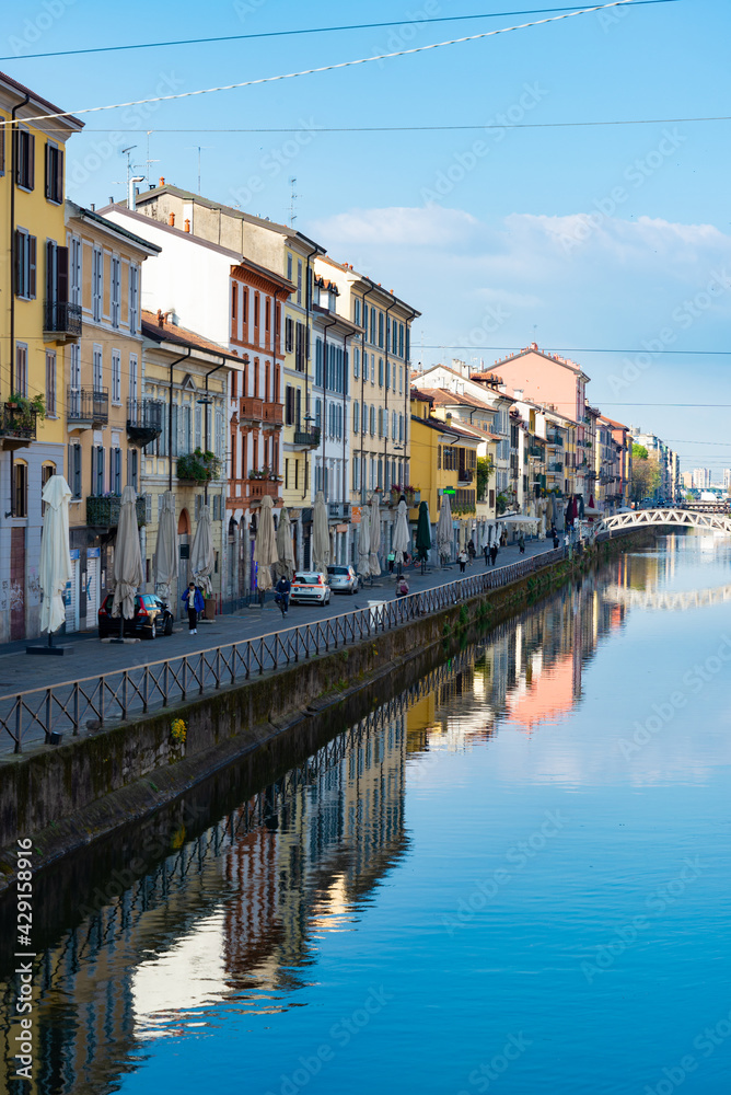 Perspective of the famous old Navigli neighborhood, Milan, Italy. Water canal in the foreground, looking like a river. Buildings and blue sky in the background.