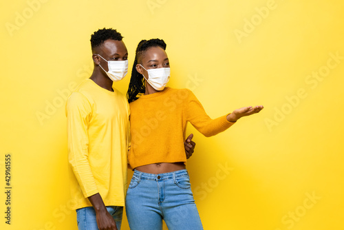 Young hygienic African couple wearing face mask protecting from Coronavirus and opening hand to copy space aside in yellow isolated studio background