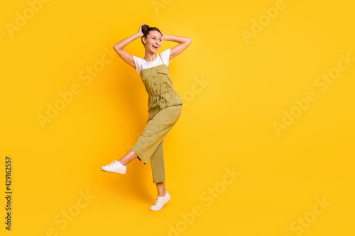 Full size photo of optimistic brunette lady dance look empty space wear bright t-shirt overall sneakers isolated on yellow color background