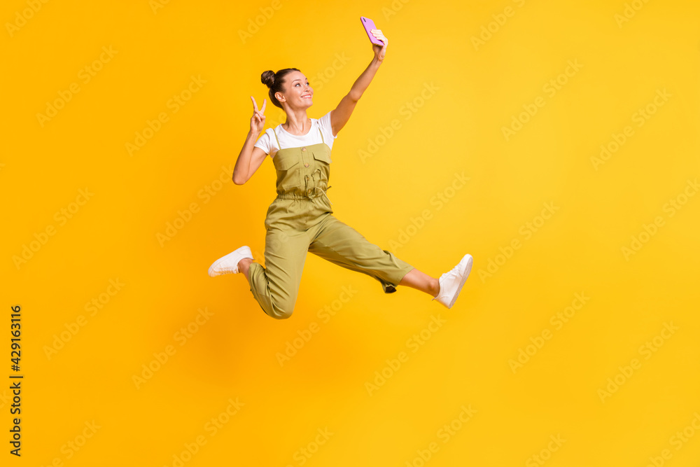 Photo of funky lady jump hold phone make selfie show v-sign wear green overall sneakers isolated yellow background