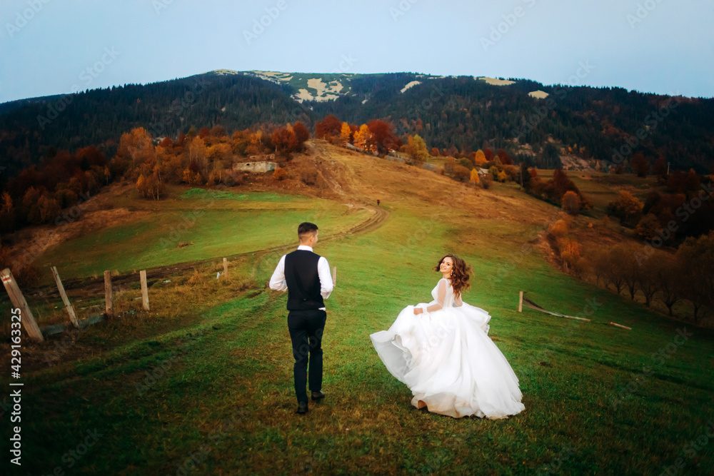 Pretty couple of newlyweds man groom and woman bride in wedding dresses run against the background of mountains Pretty couple of newlyweds man groom and woman bride in wedding dresses run against the background of mountains