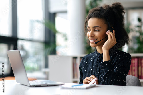 African American friendly young woman with headset, call center worker, consultant, business person, looks at the laptop screen, talking with colleagues or clients by video call, conducts consultation photo