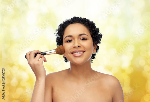 beauty, cosmetics and people concept - portrait of happy smiling young african american woman with make up brush applying blush to her face over yellow lights background