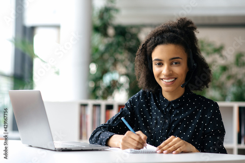 Charming successful African American young woman, in a headset, freelancer, employee or manager, sits at a work table with a laptop and a notebook, takes notes, looks at the camera, smiles friendly