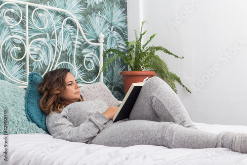 woman lying in bed reading a book, brunette woman in gray pajamas reads a book lying on a bed with a white coverlet, next to the bed there is a fern